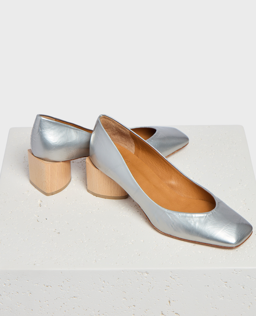 A pair of sleek, metallic silver leather shoes with a square toe design, featuring a low, chunky wooden heel and a smooth, tan interior lining.