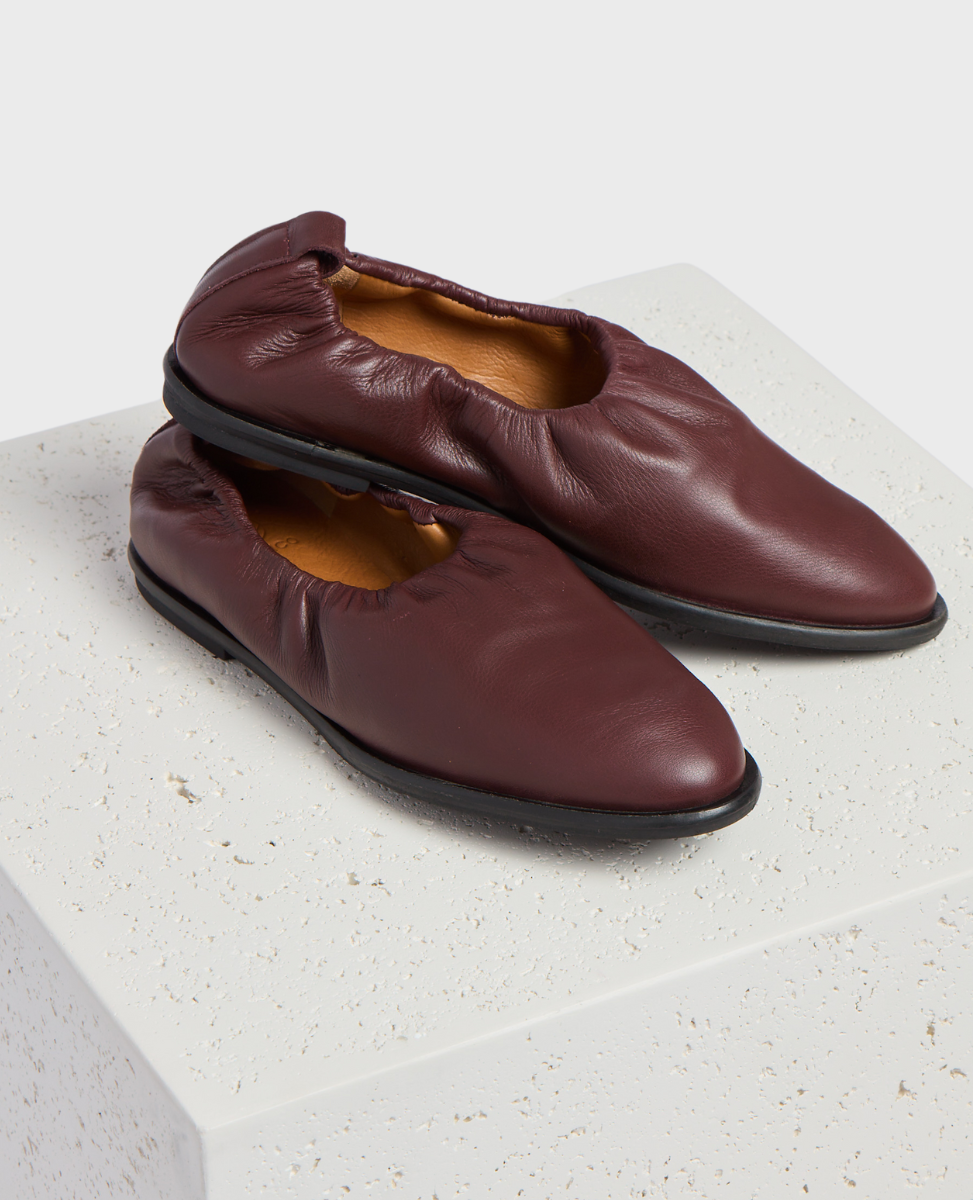 A pair of burgundy leather slip-on shoes with a soft, gathered texture, featuring a rounded toe and a black sole, resting on a textured white surface.