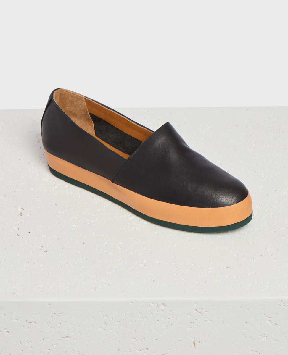 A sleek, black leather slip-on shoe with a smooth texture, featuring a contrasting tan rubber sole and a minimalist design.
