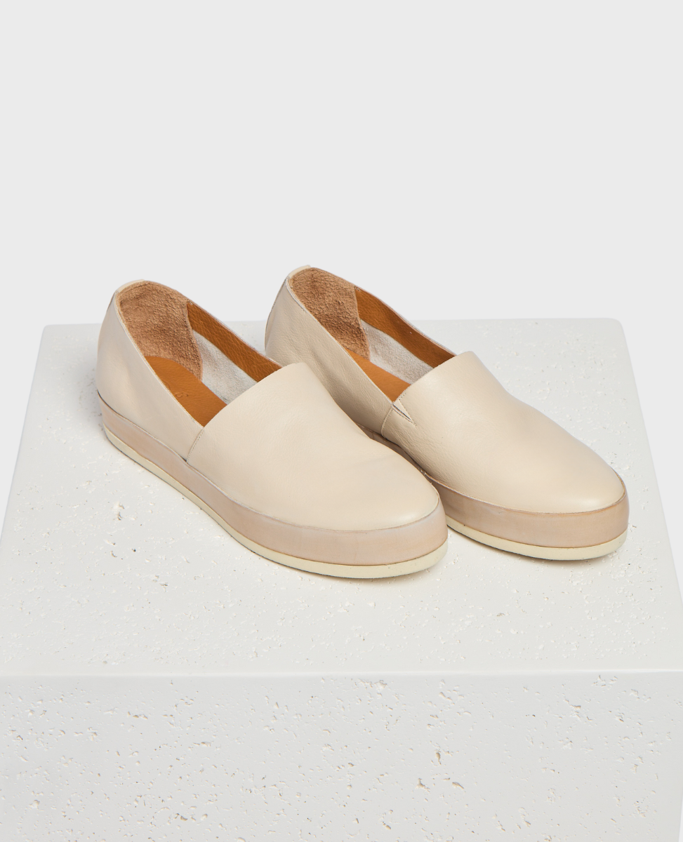 A pair of beige slip-on shoes with a smooth leather upper, rounded toe, and a flat, slightly elevated sole, featuring a minimalist design and a soft, textured interior lining.