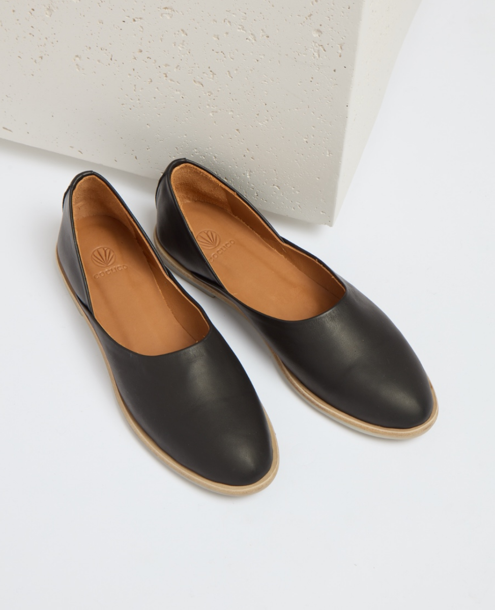 A pair of sleek, black leather slip-on shoes with a smooth texture, almond-shaped toe, tan interior lining, and a light brown flat sole.
