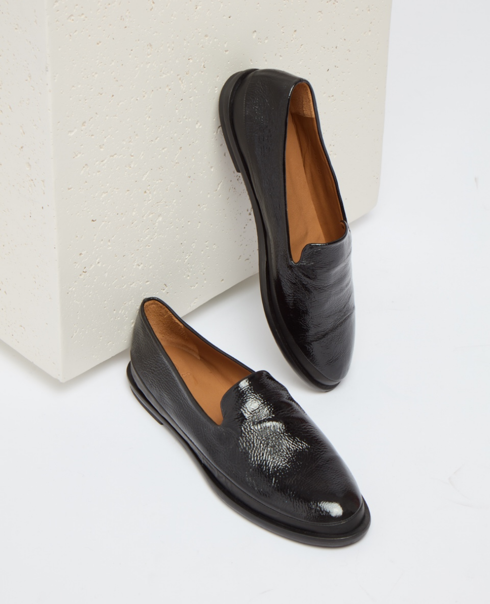 Sleek black leather loafers with a glossy finish, featuring a low heel, rounded toe, and tan interior lining, positioned against a textured white backdrop.