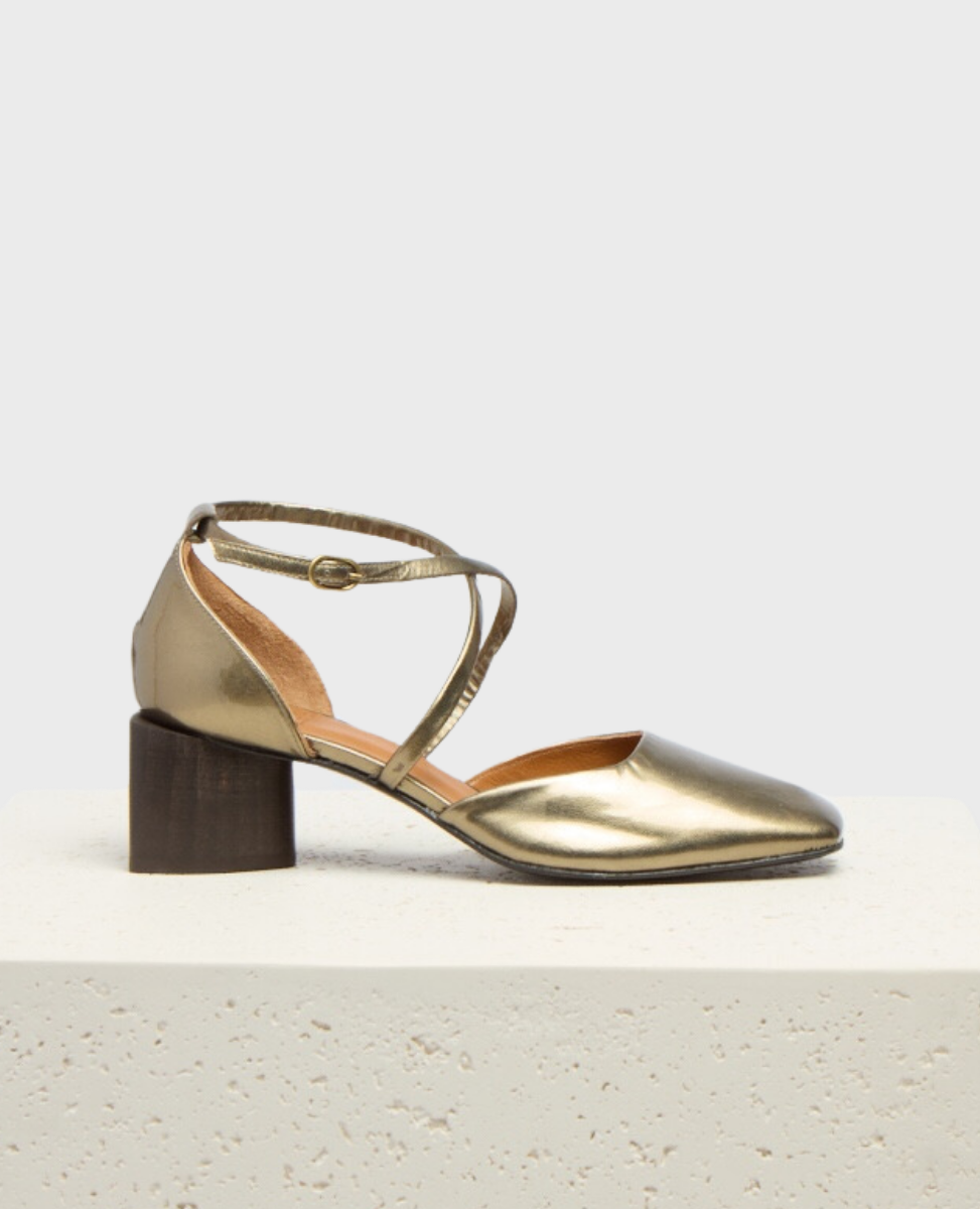 A metallic gold, closed-toe shoe with a low, chunky wooden heel and a crisscross ankle strap featuring a small buckle.