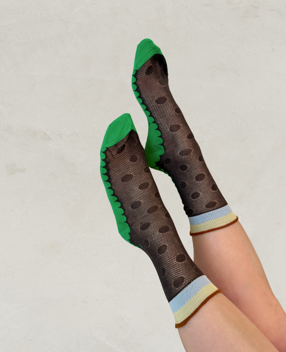A pair of socks featuring a sheer black mesh with large polka dots, green toe and heel caps, and a ribbed cuff with blue and beige stripes.