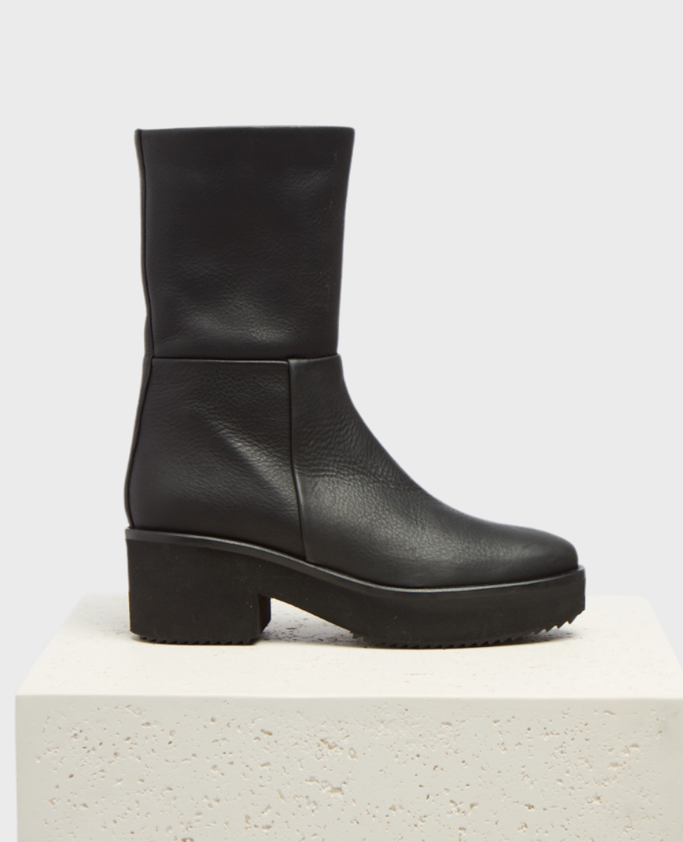 A sleek black leather boot with a mid-calf height, featuring a smooth, matte texture, a rounded toe, and a chunky block heel on a sturdy rubber sole.