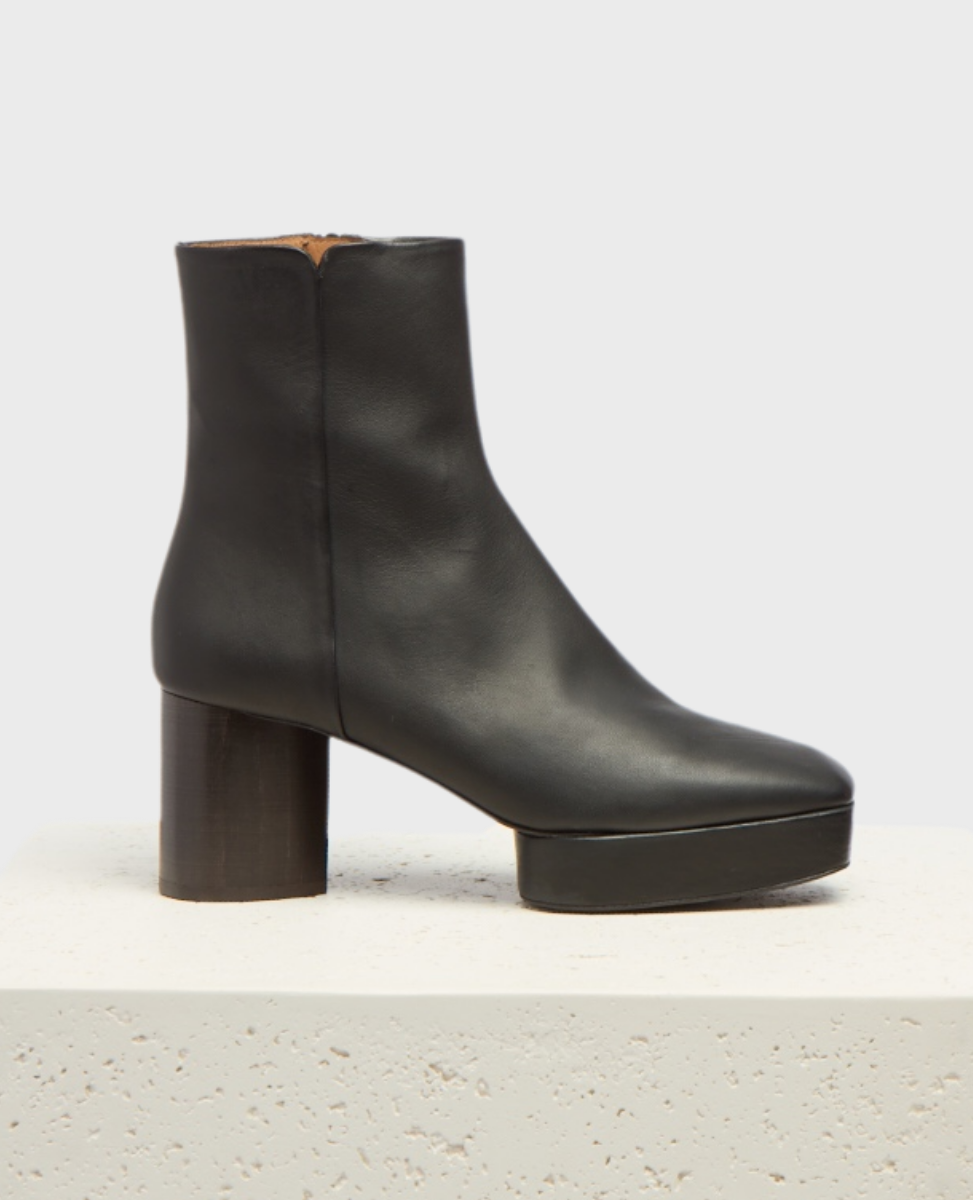 A sleek black leather ankle boot with a smooth texture, featuring a rounded toe, a chunky cylindrical heel, and a subtle platform sole.