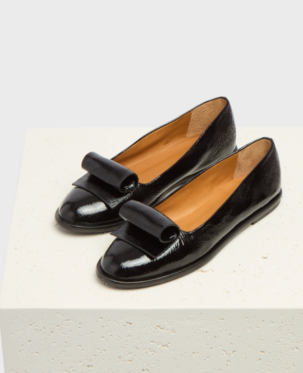 A pair of sleek, black patent leather loafers with a glossy finish, featuring a distinctive folded strap across the top, a rounded toe, and a tan interior lining, resting on a textured white surface.