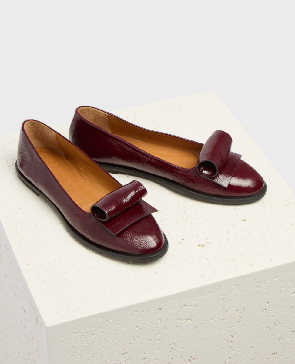 A pair of burgundy leather loafers with a glossy finish, featuring a distinctive looped strap detail on the front, a rounded toe, and a low black sole, displayed on a textured white cube.