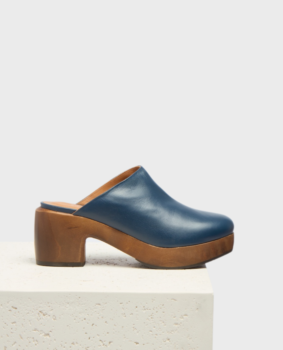 A blue leather clog with a smooth texture, featuring a rounded toe and a chunky wooden heel and platform, resting on a light-colored textured surface.