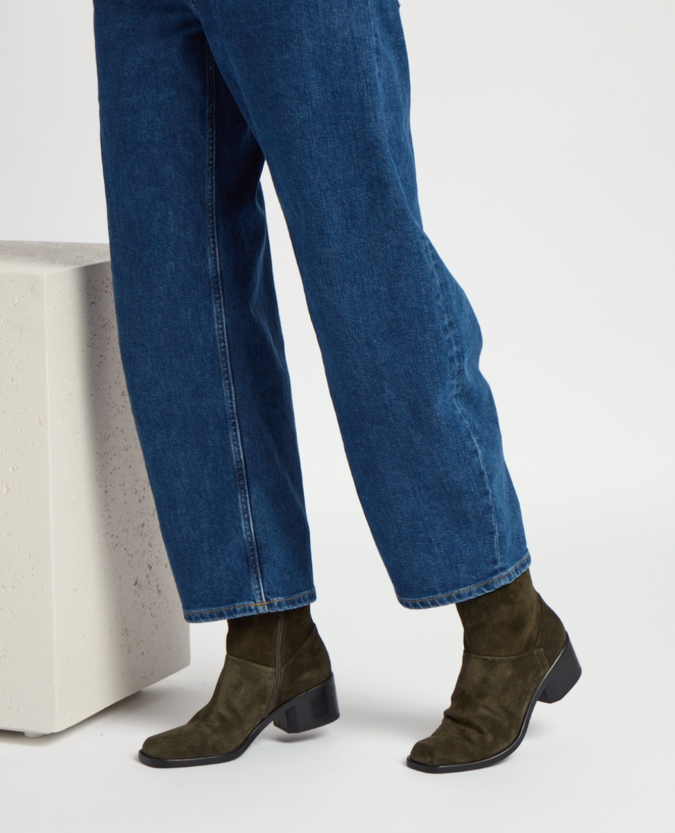 Dark olive green suede ankle boots with a block heel, featuring a sleek, minimalist design and a sqauare toe, paired with blue denim jeans.