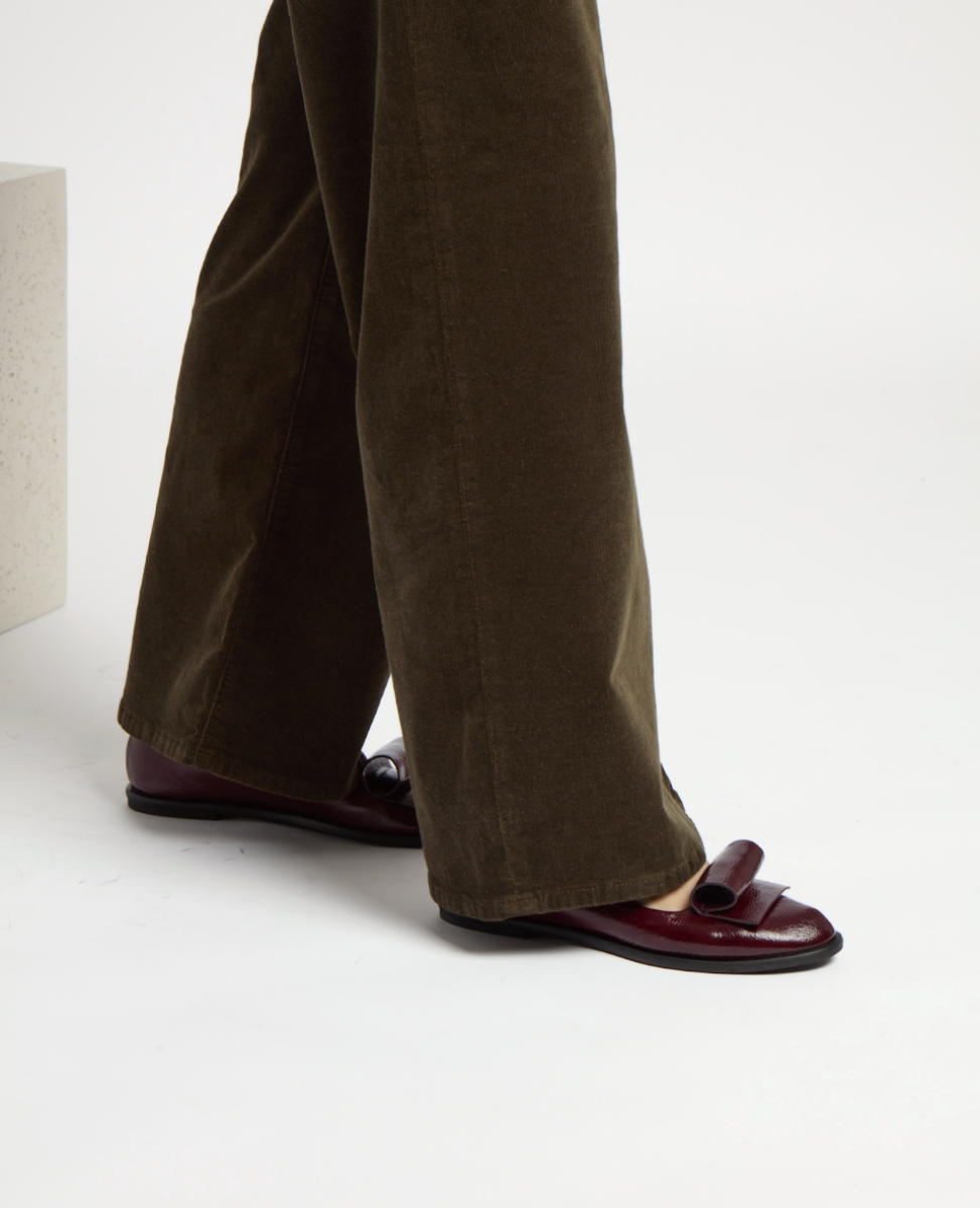 A pair of burgundy leather loafers with a glossy finish, featuring a rounded toe and a decorative strap across the top, worn with brown corduroy pants.