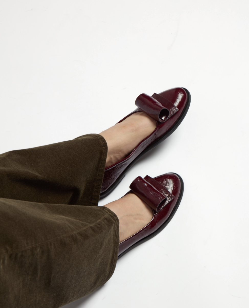 Shiny burgundy loafers with a sleek, rounded toe and a decorative strap across the top, featuring a low black sole and a smooth, polished leather finish.
