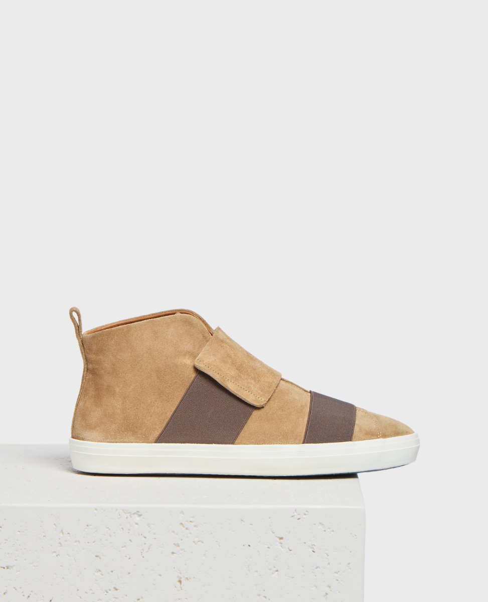 A tan suede high-top sneaker with a white rubber sole, featuring a wide brown elastic band across the midfoot and a pull tab at the heel.