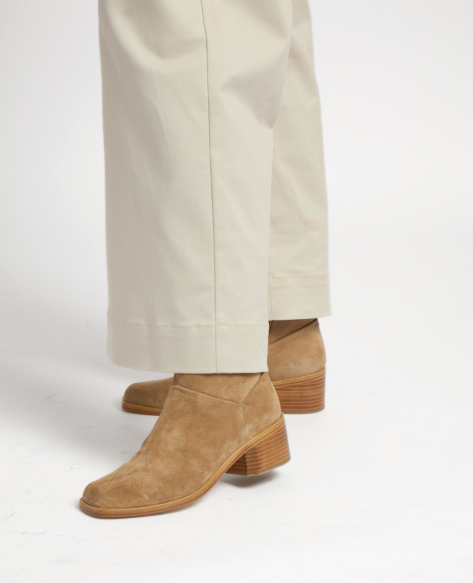 Tan suede boots with a block heel and squared toe, featuring a smooth texture and a wooden sole, paired with cream-colored wide-leg pants.