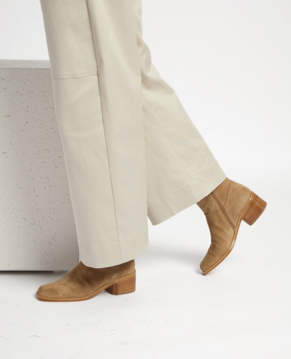 A pair of tan suede boots with a low wooden block heel, featuring a smooth texture and a sleek, minimalist design, worn with light beige wide-leg pants.
