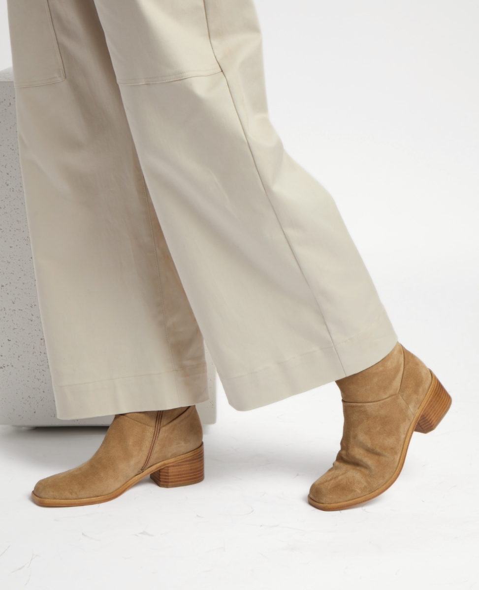 Tan suede boots with a squared toe, wooden block heel, and subtle stitching details, paired with wide-leg cream pants.