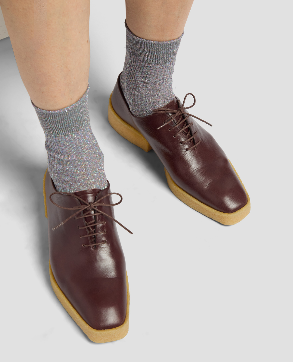 Brown leather shoes with yellow soles worn with gray socks on a light gray background