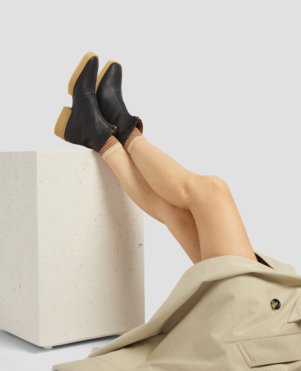 Person wearing black ankle boots with tan soles, sitting on a white box against a light gray background.