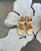 The Sterling wood heeled, strappy sandal in champagne metallic suede, shown on a wood surface 3