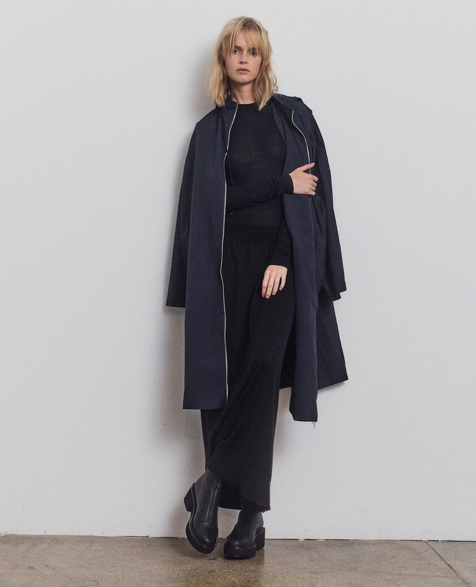 Person wearing a long navy coat over a black outfit against a plain white wall.