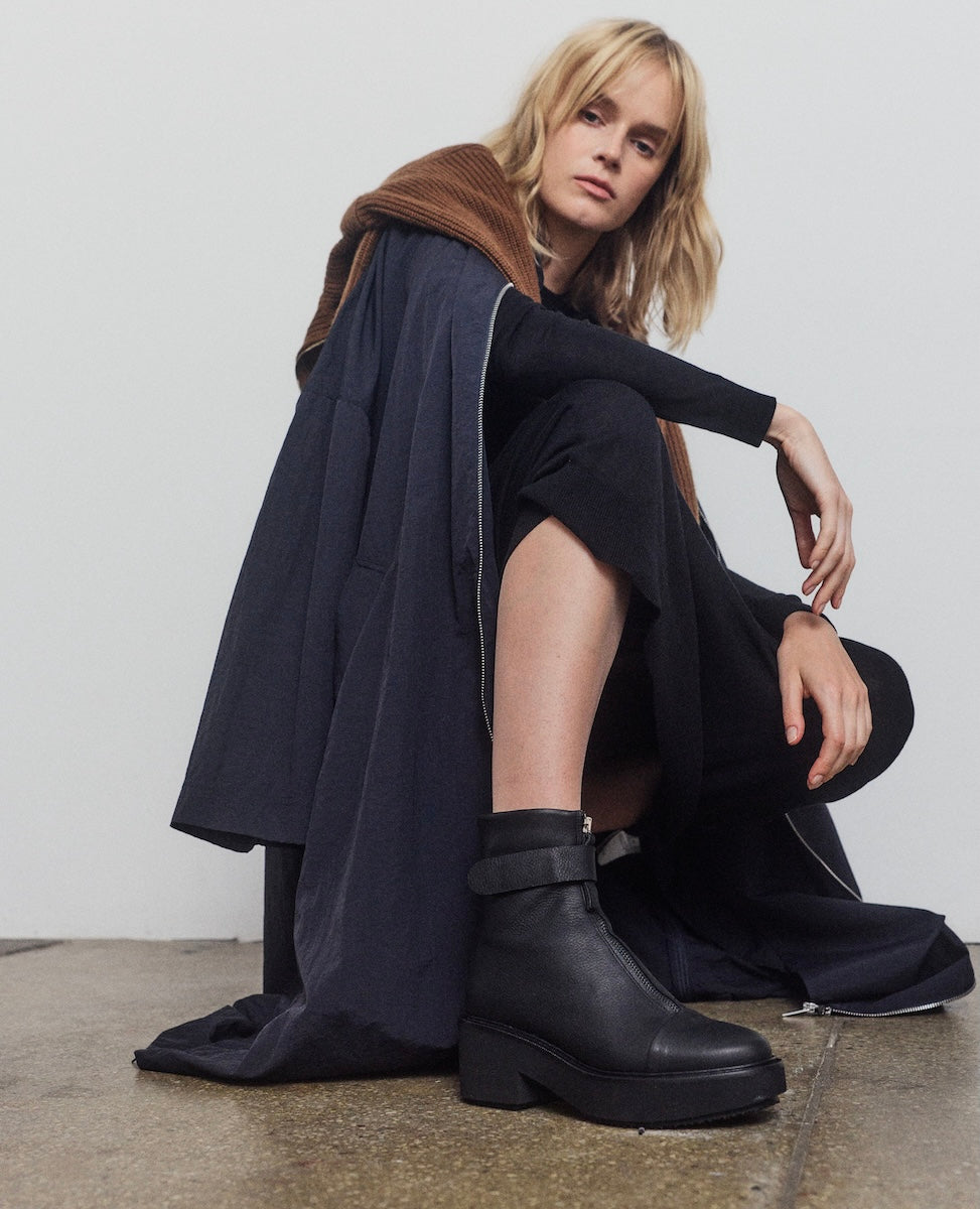 Woman wearing a black coat and boots sitting on the floor against a white wall.