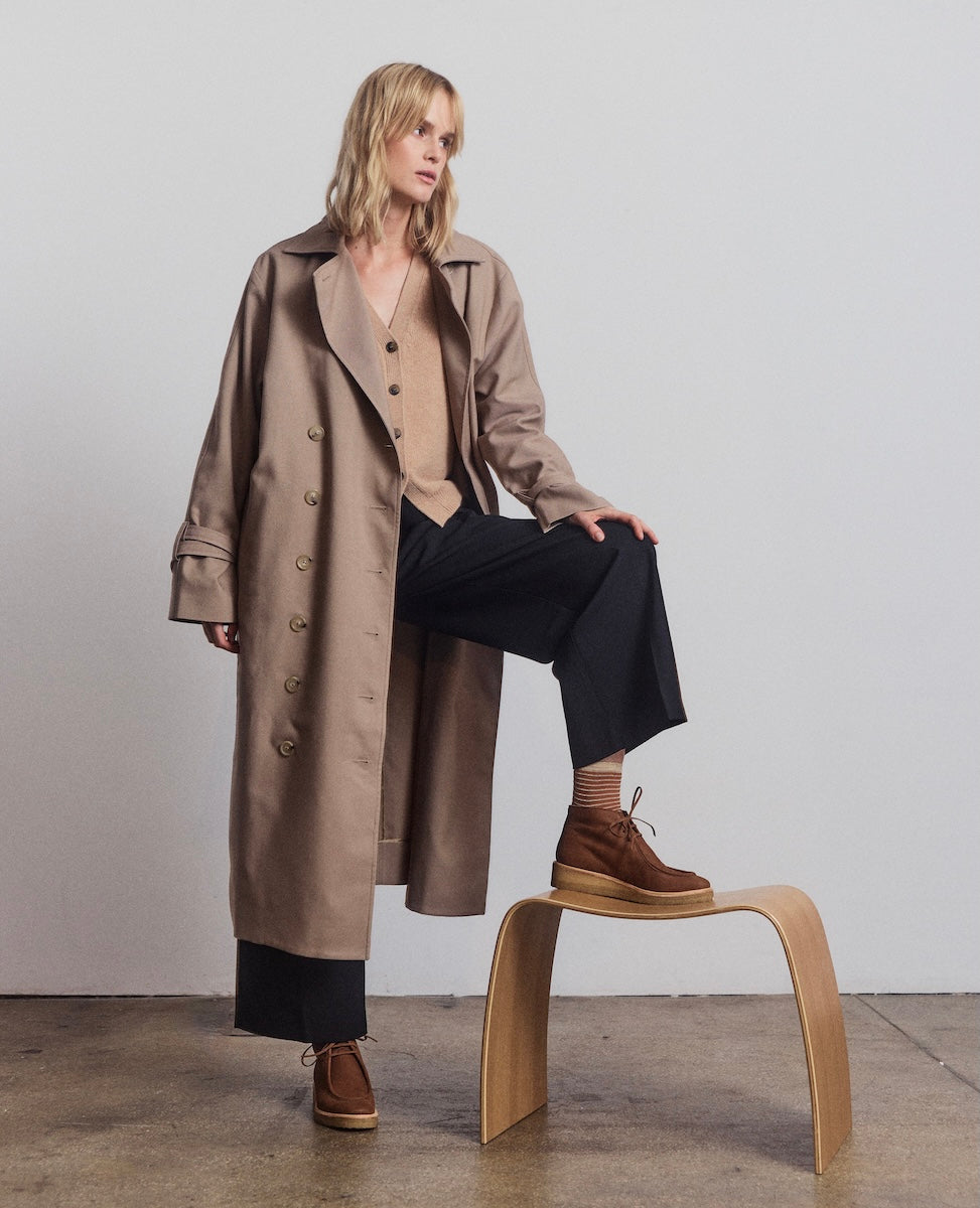 Woman wearing a long brown coat sitting on a wooden stool against a plain background