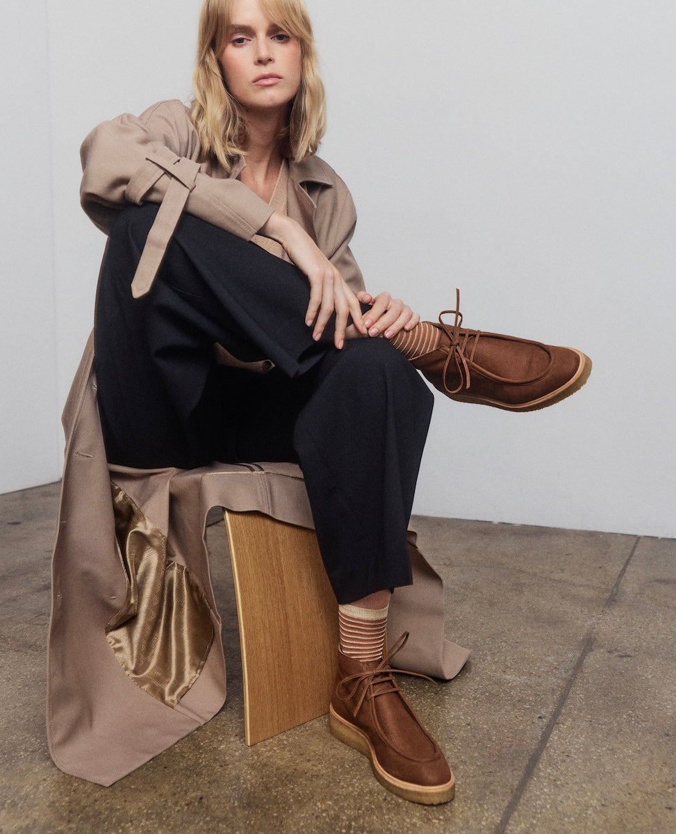 Woman sitting on a wooden stool wearing a beige trench coat, black outfit, and brown shoes.