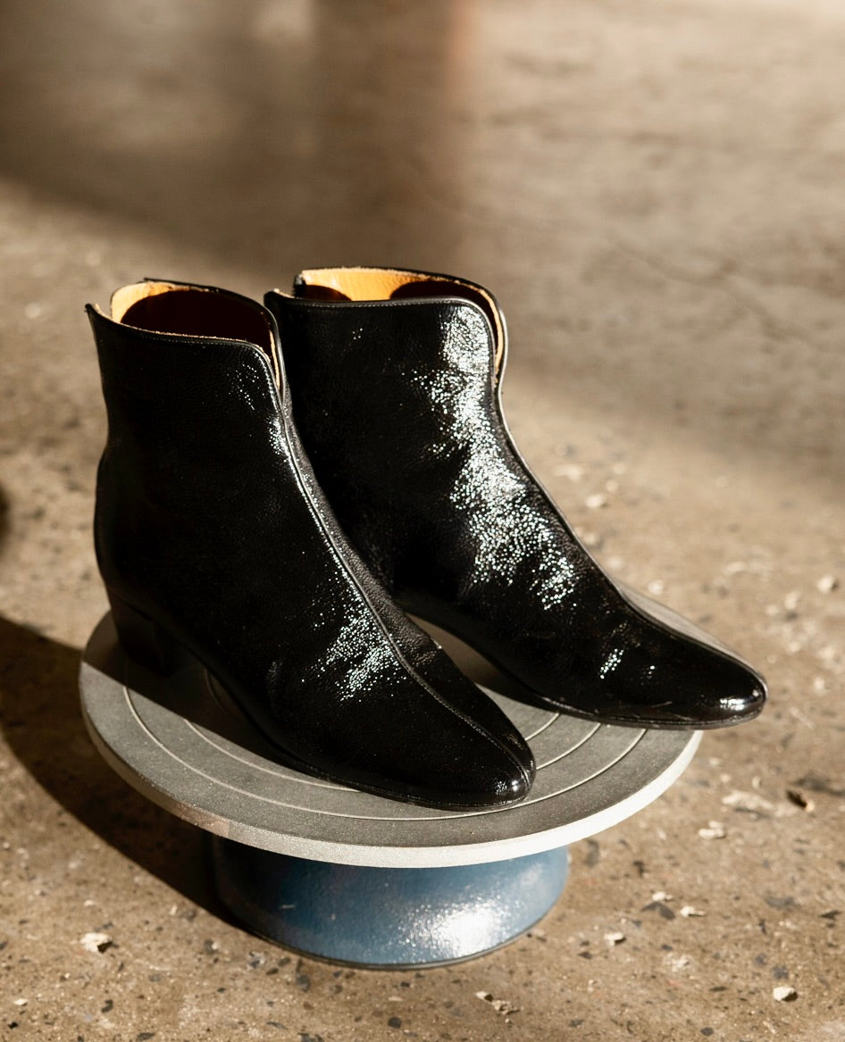 A pair of sleek, black, patent leather ankle boots with a glossy finish, almond shaped toes, and a low wood block heel, displayed on a round, gray pedestal.