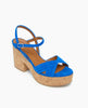 A vibrant blue suede sandal features a chunky cork platform heel, crisscross straps over the toe, an adjustable ankle strap with a small buckle, and a smooth tan insole. 5