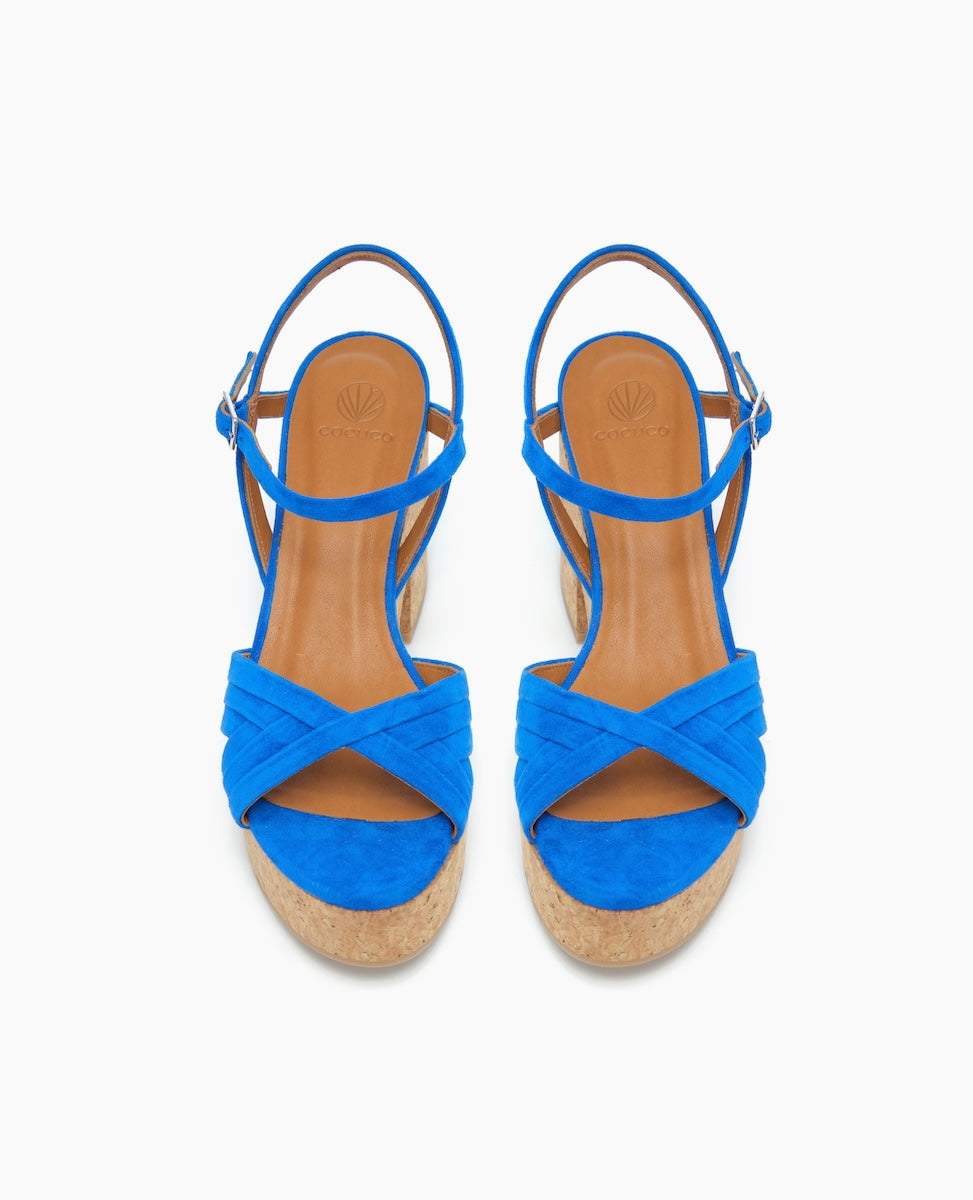 Bright blue sandals with crisscross suede straps, a tan leather insole, adjustable ankle straps, and a cork wedge heel.