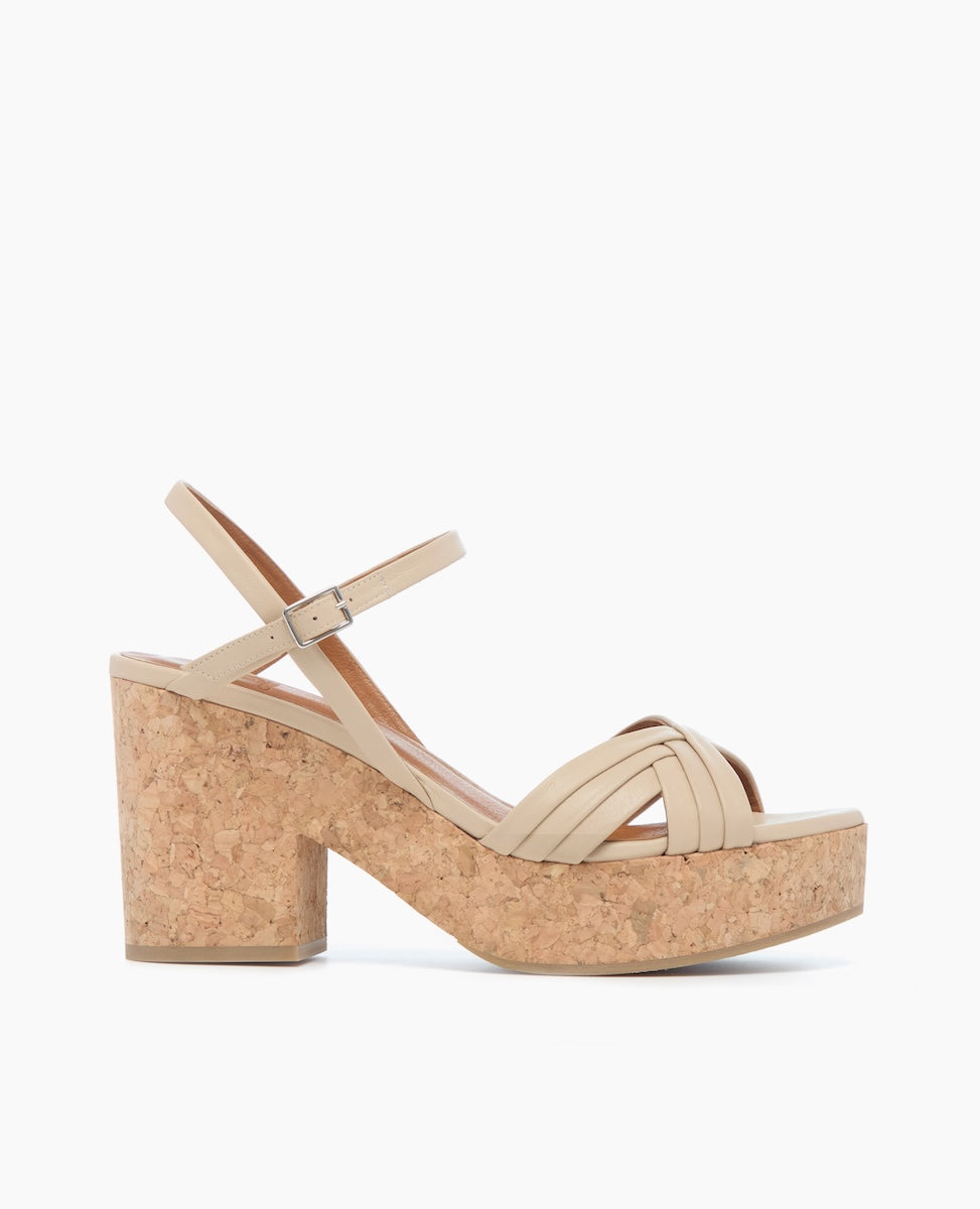 A beige sandal with a cork platform heel features crisscrossing straps over the toe, an adjustable ankle strap with a silver buckle, and a chunky, textured heel.