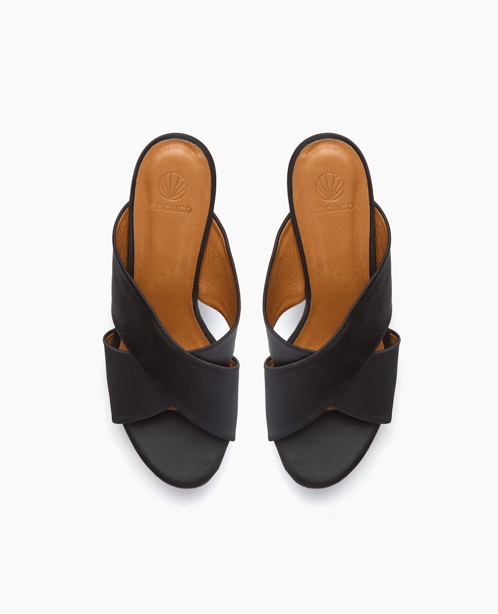 A pair of black, open-toe sandals with wide crisscross straps, featuring a tan leather insole and a subtle logo embossed near the heel.