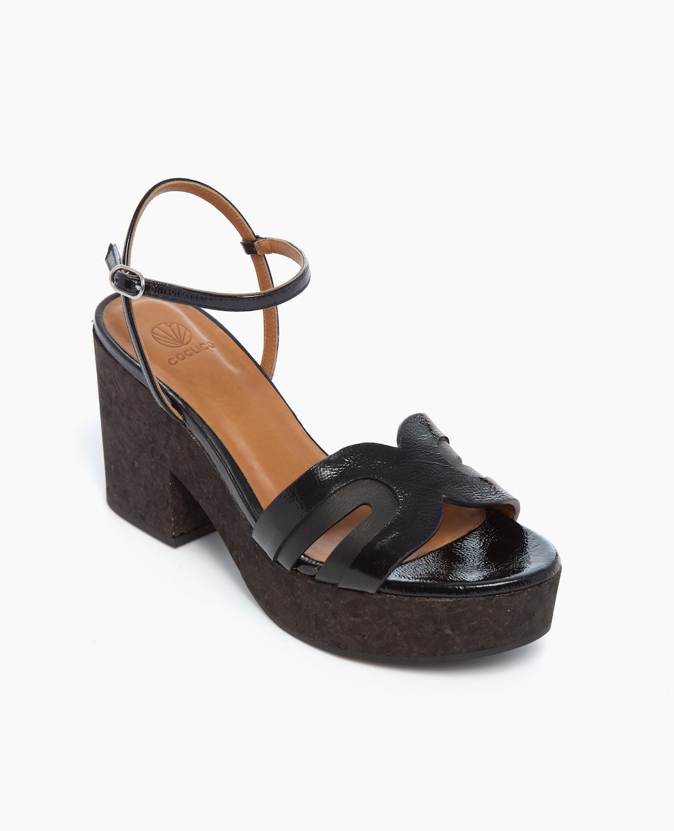 A black, open-toe platform sandal with a thick, textured heel, featuring a wavy strap design and an adjustable ankle strap, complemented by a tan insole.