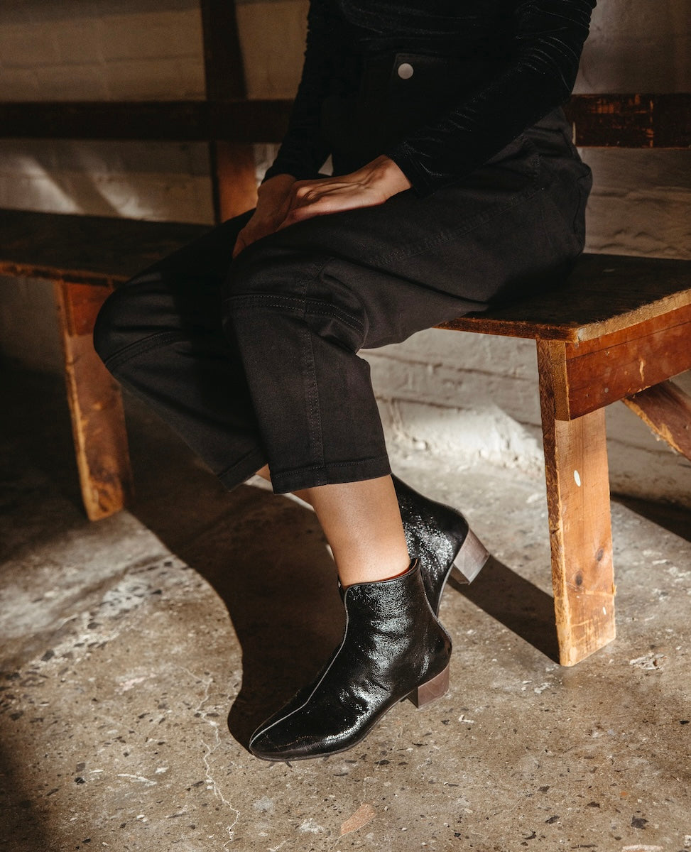 A person is seated on a wooden bench, wearing black ankle boots with a glossy finish, an almond shaped toe, and a low, solid wood heel, paired with black pants.