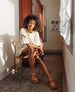 Woman sitting on a wooden chair in a white sequin top and the Dana Wedge in Savana Cuoio.  2