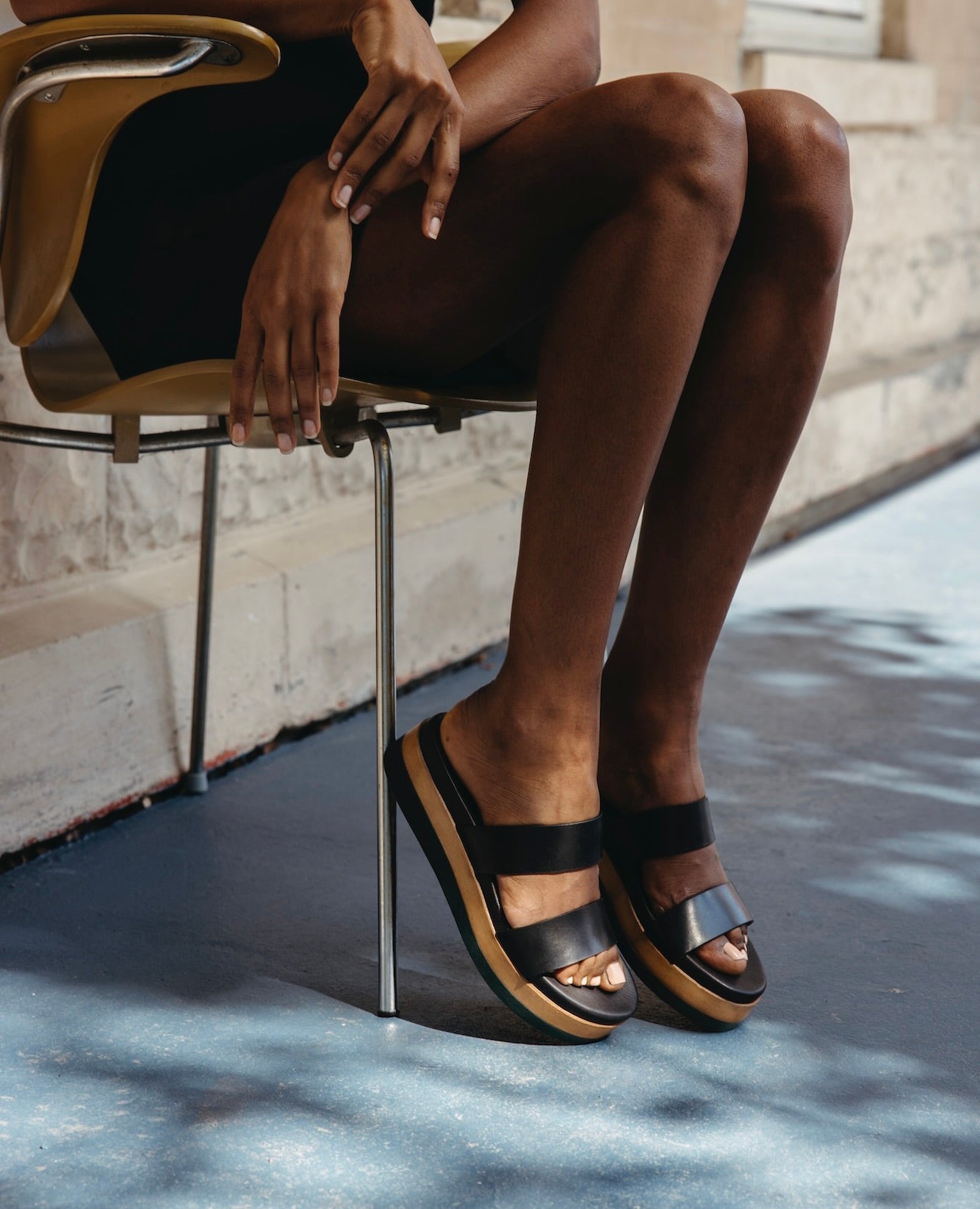 Legs of a woman sitting on a wooden chair with her arms leaned over the legs and the toes pointed down.