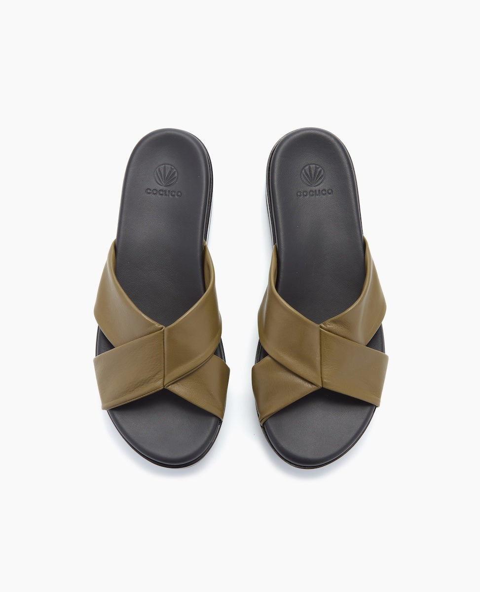 A pair of olive green slide sandals with wide, crisscrossing straps over a black footbed, featuring a smooth, matte texture and a subtle logo embossed on the insole.