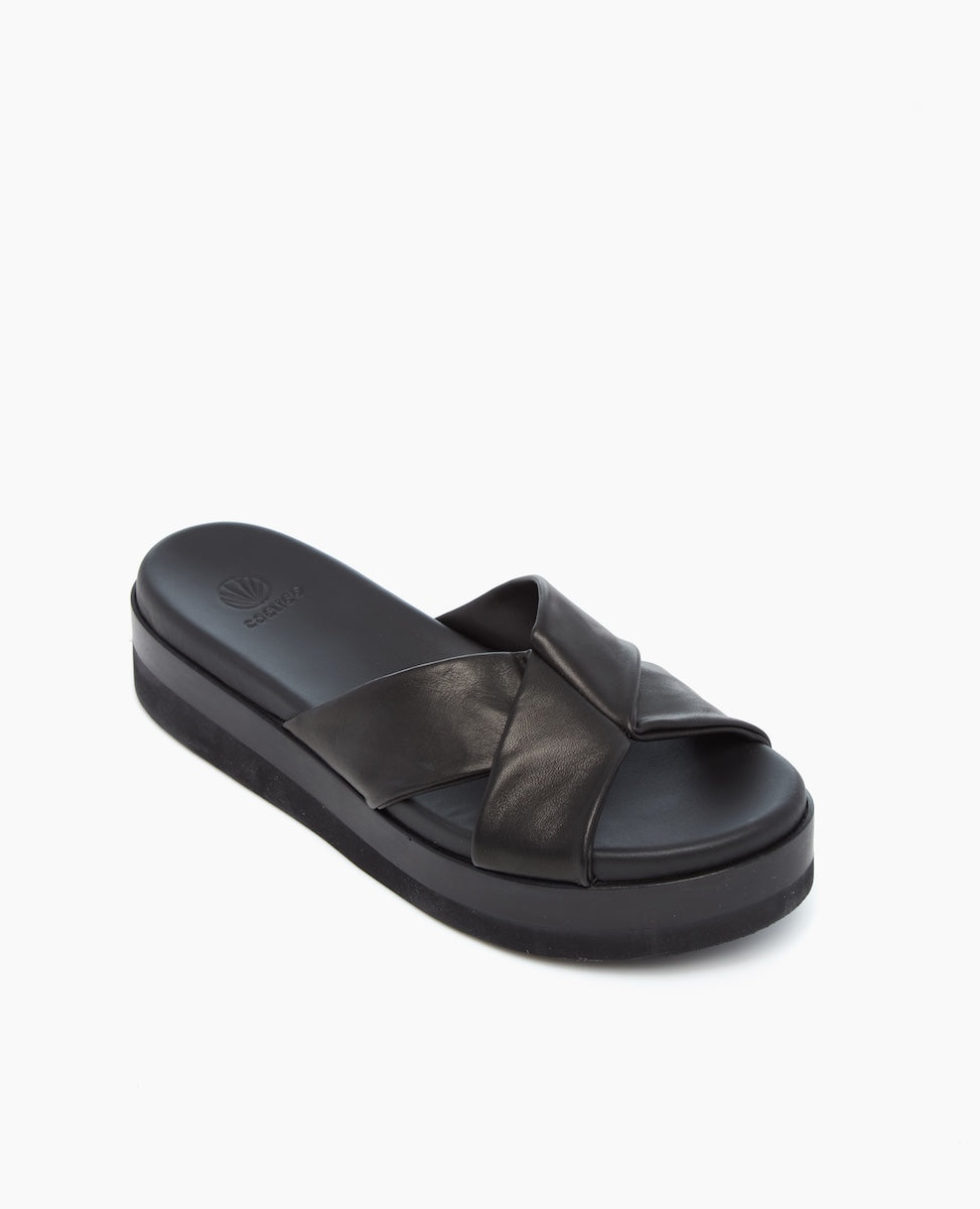 A black slide sandal featuring a thick, cushioned sole and wide, crisscross leather straps with a smooth texture.