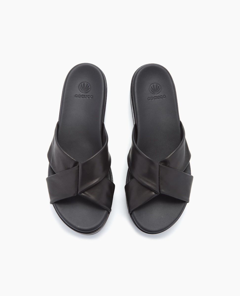 A pair of sleek black slide sandals featuring a crisscross strap design, smooth leather texture, and a subtle logo embossed on the footbed.