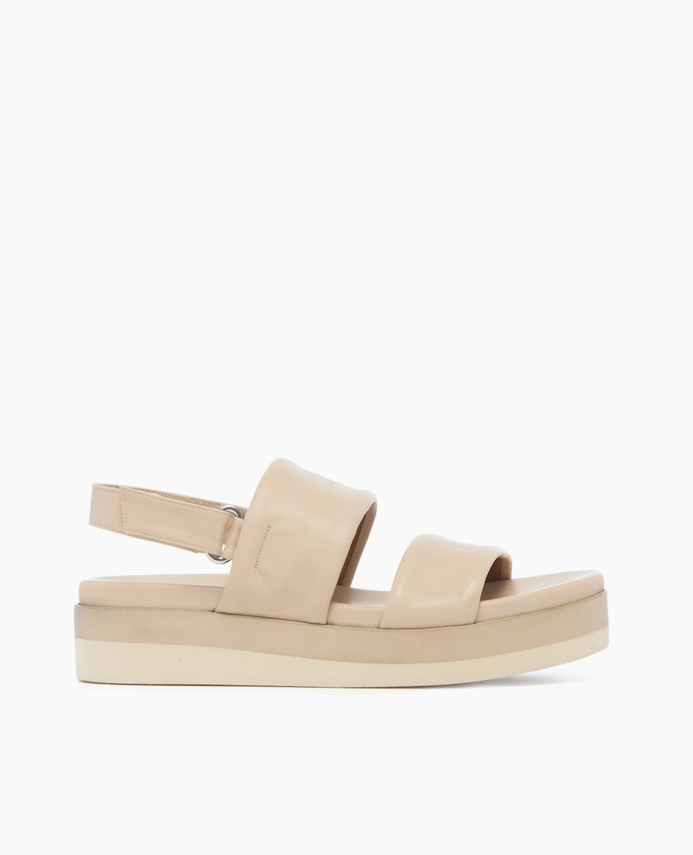 A beige sandal with a flat, thick sole, featuring a wide strap across the front and an adjustable ankle strap, made of smooth leather material.