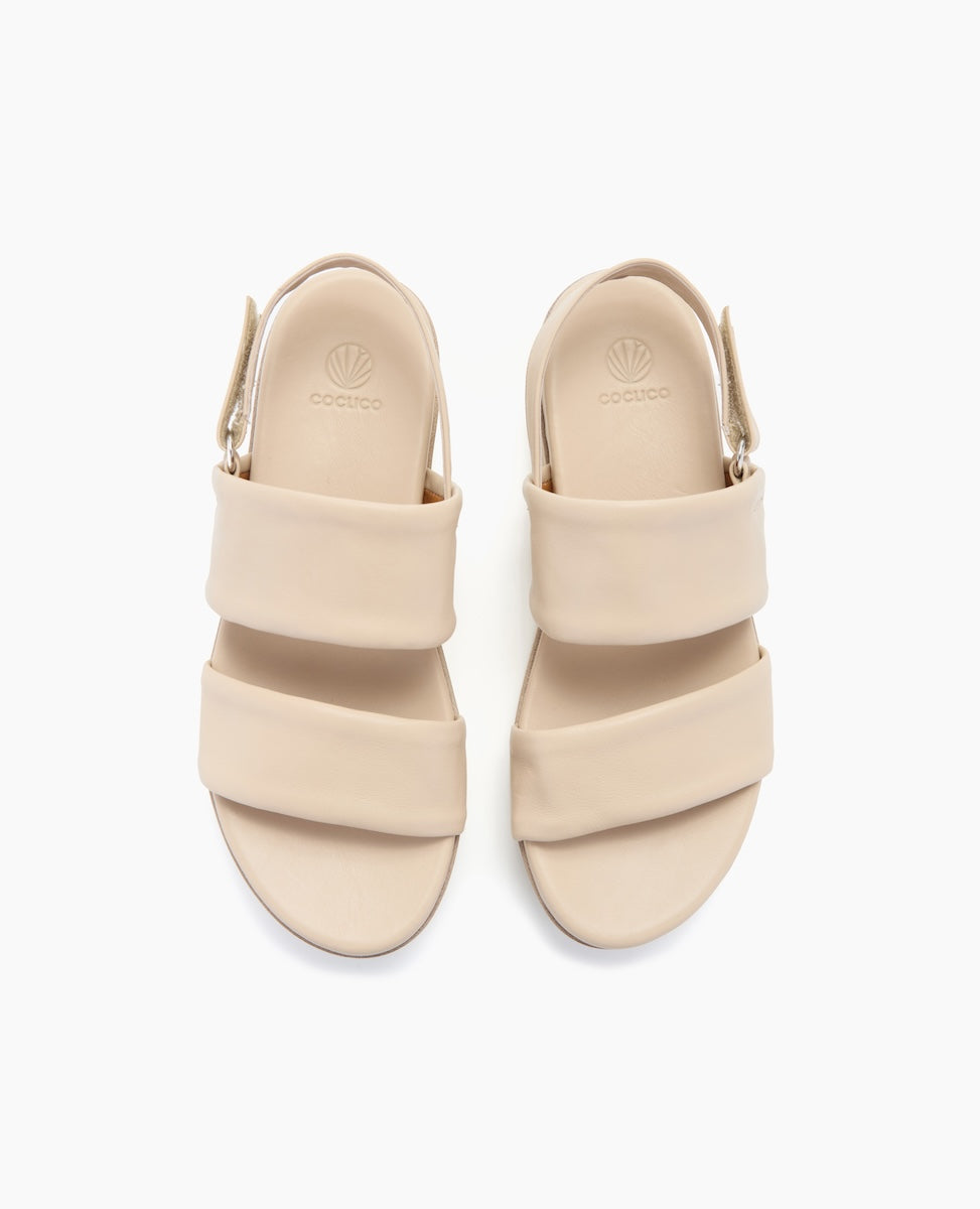 Beige sandals with a smooth, matte finish feature two wide straps across the top, a rounded open toe, a slingback strap with a buckle, and a slightly elevated sole.