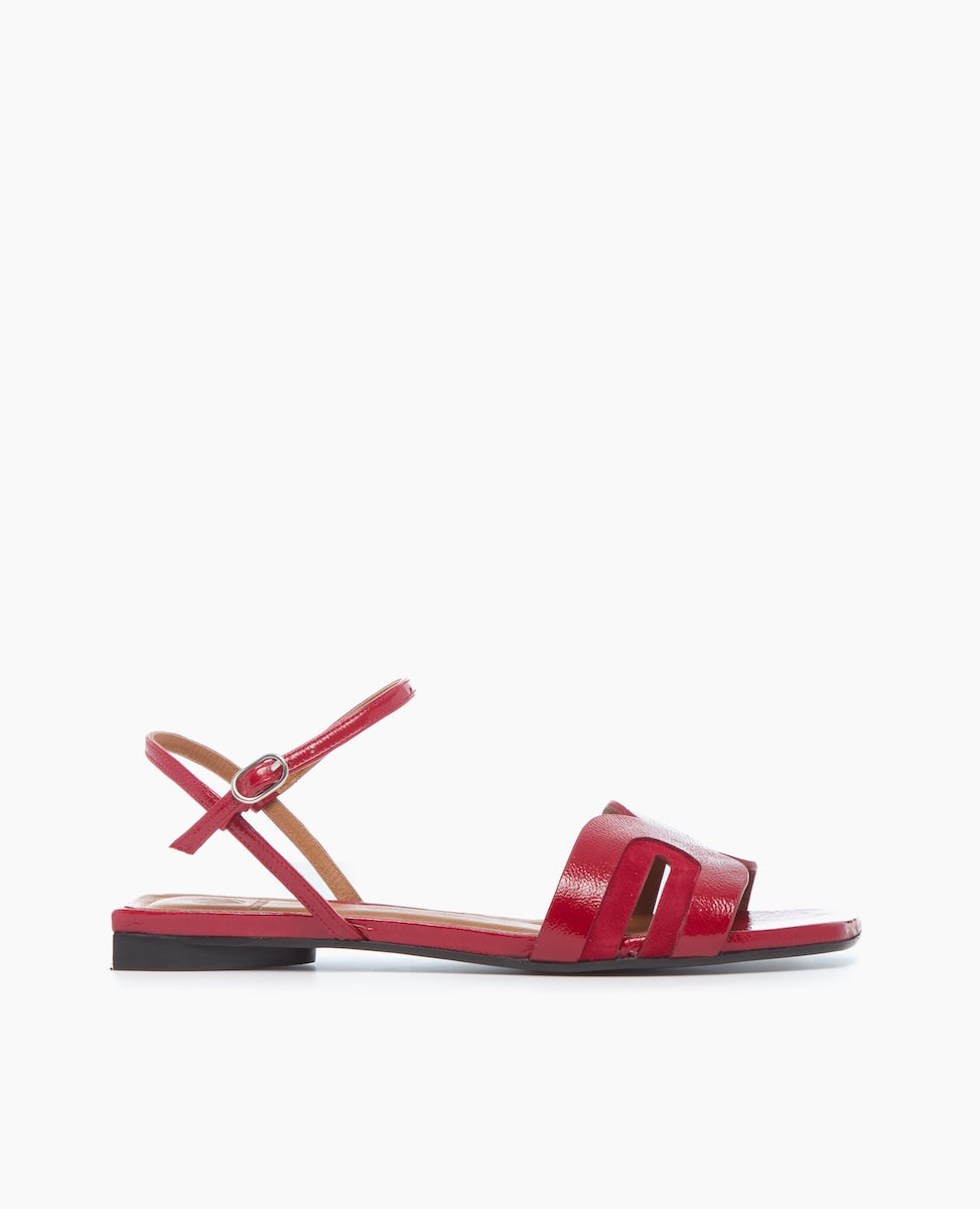 A sleek red sandal with a glossy finish features a flat sole, an open toe design, a thin ankle strap with a buckle, and a distinctive cut-out pattern on the front strap.