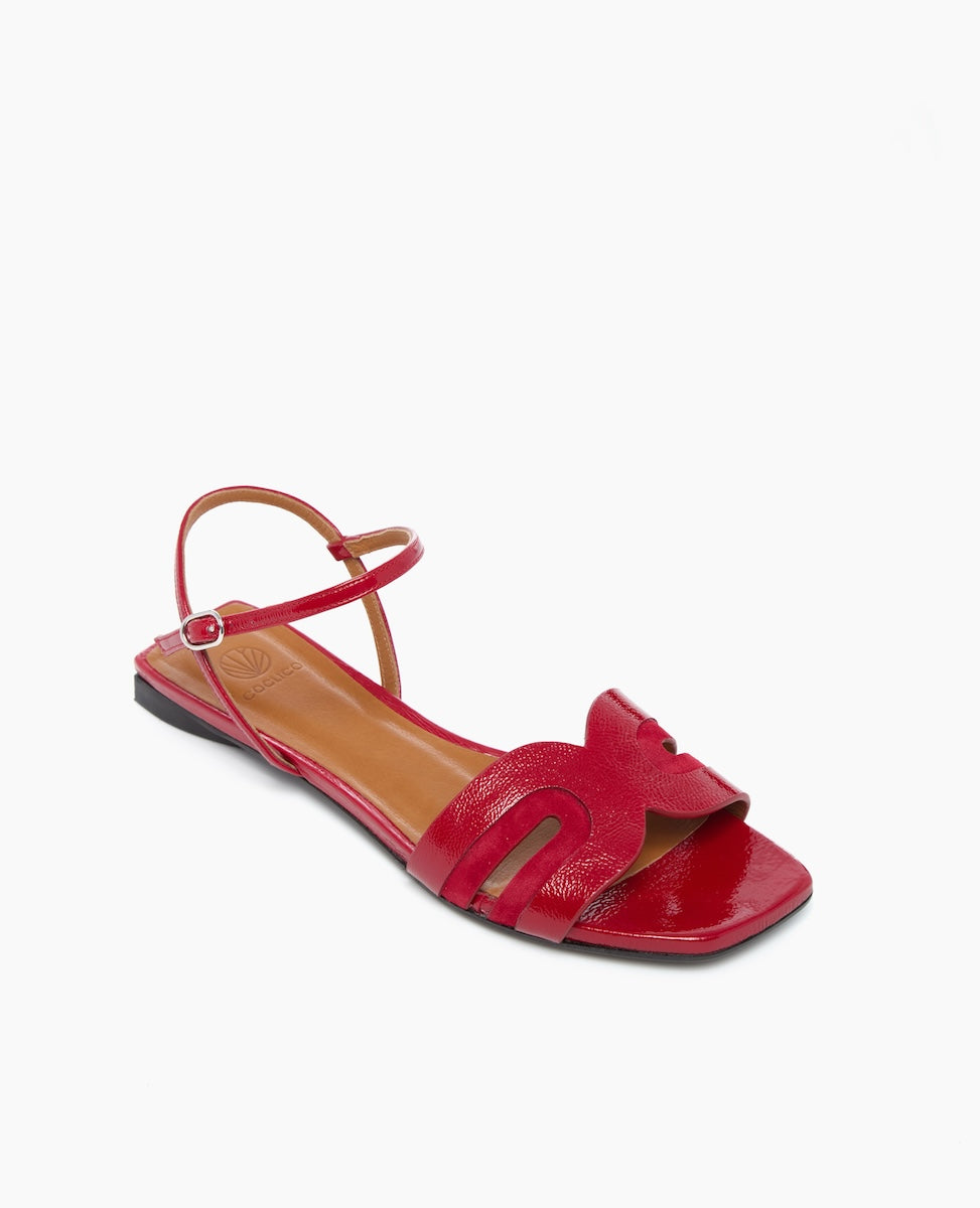 A vibrant red sandal with a glossy finish features a flat sole, an open toe design with a wavy cutout pattern, a thin ankle strap, and a small buckle closure, crafted from smooth leather.