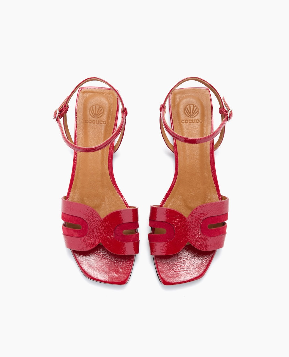 Red sandals with a glossy finish feature a unique cut-out design on the front strap, an adjustable ankle strap, and a tan insole with a logo imprint.