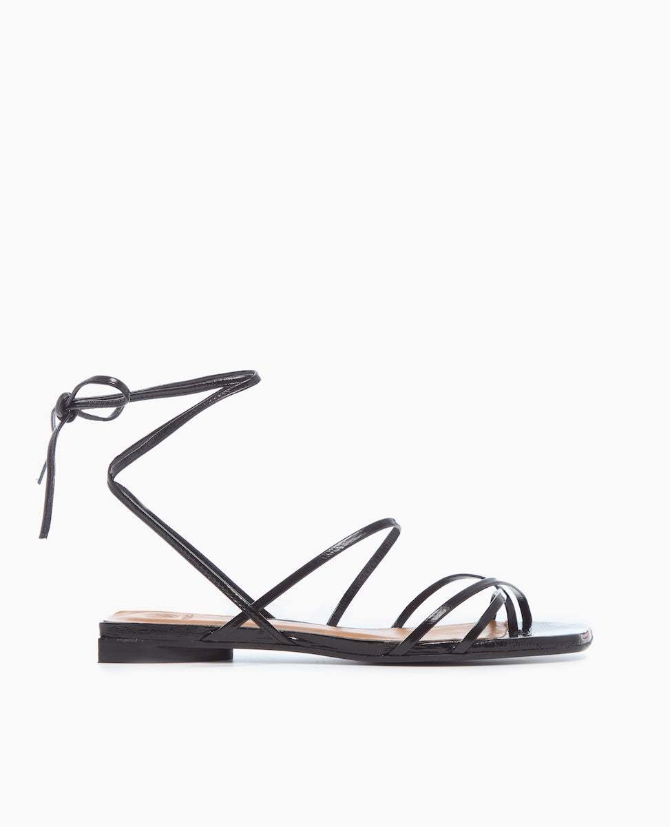 A sleek black sandal featuring thin, glossy straps that wrap around the ankle, a flat sole, and a minimalist design with a knot detail at the back.