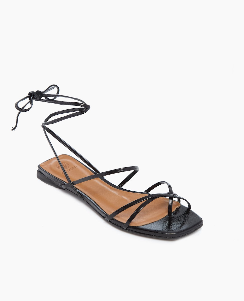 A sleek black sandal featuring thin, glossy straps that crisscross over the foot and wrap around the ankle, with a flat sole and a square toe design.