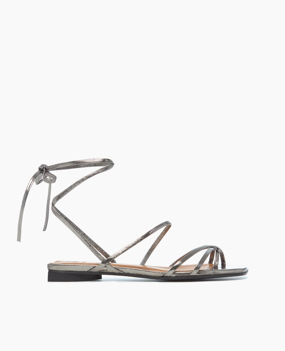 A sleek, metallic silver sandal with thin, crisscrossing straps and a delicate ankle tie, featuring a flat sole and minimalist design.