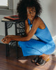 Woman wearing a blue dress bent down wearing the Ferhana Sandal in anthracite shimmer suede. 5