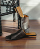 Close up view of the Ferhana Sandal in anthracite shimmer sued with the heel of the left sandal propped up against the night table. 9