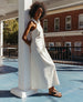 Woman wearing a white dress leaning against a white column wearing the Finch Sandal in Black.  7